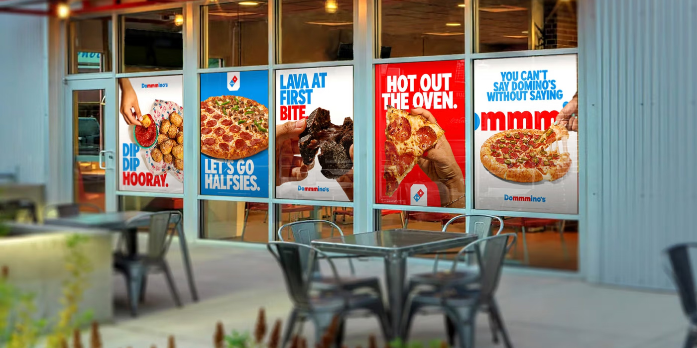Restaurant window with colorful Domino's ads featuring pizza and desserts. Outdoor seating area with metal tables and chairs; inviting and vibrant ambiance.