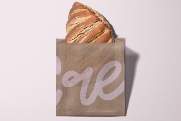 A golden-brown croissant peeks out of a rustic brown paper bag with "love" written in white script. The setting is minimalist against a light background.