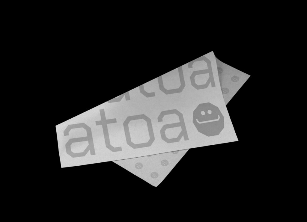 Two overlapping white papers with "ATOA" and a smiley face on them, placed against a black background. The tone is minimalist and modern.