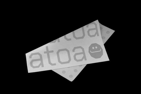 Two overlapping white papers with "ATOA" and a smiley face on them, placed against a black background. The tone is minimalist and modern.