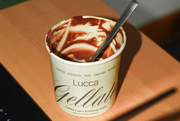 Empty ice cream cup labeled "Lucca Gelato" with chocolate remnants inside and a spoon, placed on a wooden table, casting shadows.