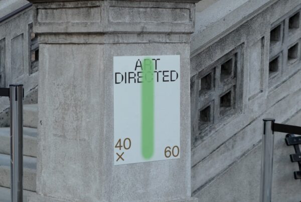 A vertical white poster with black typography and a green spray-paint detail mounted on a stone architectural pillar.