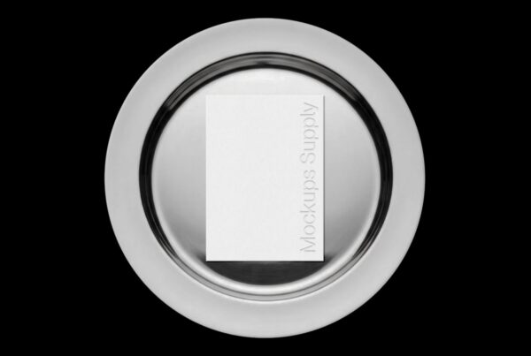 A top-down shot of a white A5 menu card with debossed text centered on a circular silver metal tray over a solid black background.
