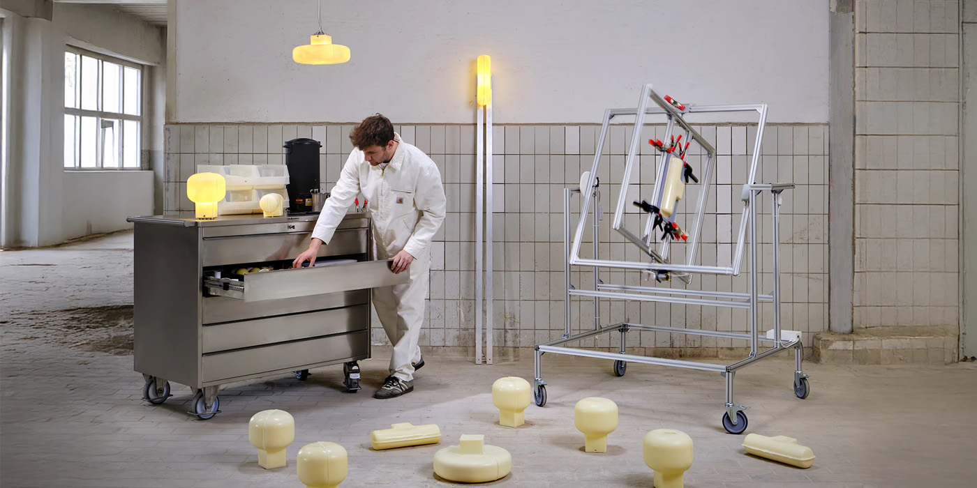 A designer in white overalls uses a stainless steel workbench while a rotational molding machine spins a sustainable wax lamp mold nearby.