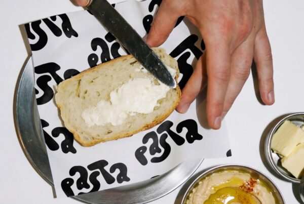 A top-down view of a hand using a knife to spread butter on a slice of bread atop branded tissue paper on a metal plate.