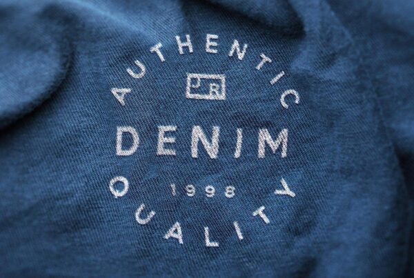 A close-up view of a circular white printed logo reading "Authentic Denim Quality" on soft blue fabric with a shallow depth of field.