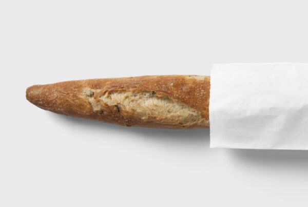 A single, fresh French baguette bread loaf extends horizontally out of a crisp white paper sleeve on a plain light background.