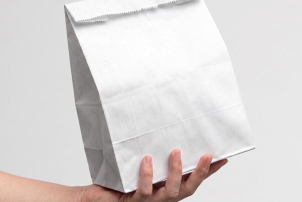 A hand holding a blank, white, folded-top paper takeaway bag against a neutral light gray background.