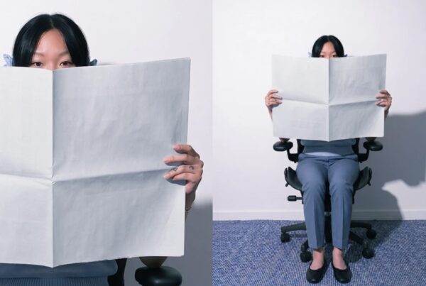 A woman sitting in an ergonomic office chair holding a folded blank broadsheet paper in front of her face against a white wall.