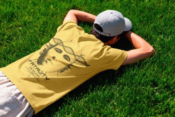 A model wearing a grey cap and a mustard yellow t-shirt with a graphic back print, lying face down on bright green grass in direct sunlight.